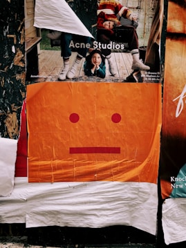 Layered posters on a wall include an advertisement featuring two people in casual, colorful clothing, with one person lying on a wooden deck. Below, an abstract poster depicts a minimalist orange face with a neutral expression, composed of simple red shapes.