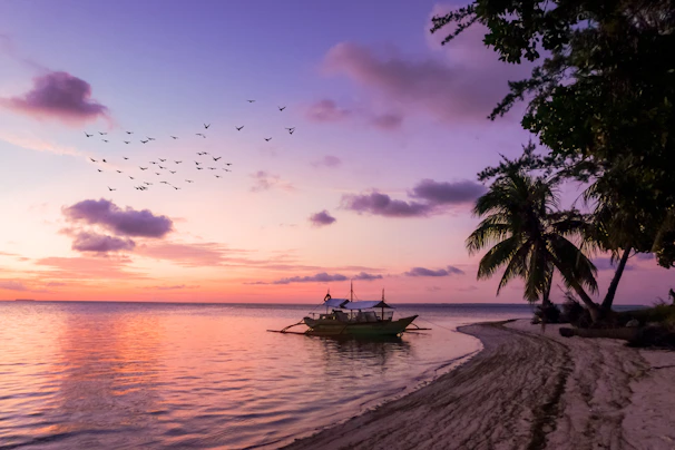 Sunset over turquoise waters with a small boat sailing near a palm-fringed shoreline.