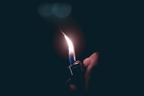 A hand is holding a metal lighter that produces a tall, steady flame against a dark background. The flame illuminates the surrounding area softly.