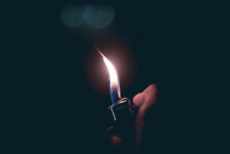 A hand is holding a metal lighter that produces a tall, steady flame against a dark background. The flame illuminates the surrounding area softly.
