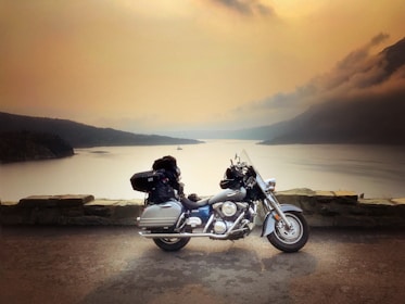 A motorcycle is parked on a concrete surface near a stone barrier, overlooking a large body of calm water and distant forested hills under a hazy sky. The vehicle is packed with luggage, suggesting travel or a road trip.
