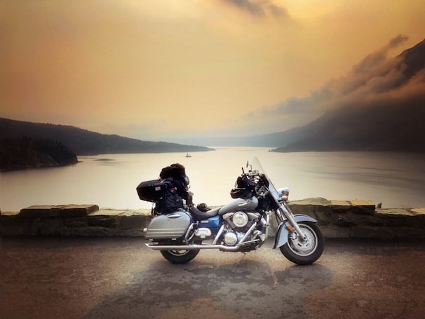 A motorcycle is parked on a concrete surface near a stone barrier, overlooking a large body of calm water and distant forested hills under a hazy sky. The vehicle is packed with luggage, suggesting travel or a road trip.