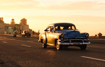 A vintage blue car drives along a coastal road during a sunset, casting warm golden hues across the scene. In the background, several older buildings are visible alongside the roadway, while a few pedestrians and another classic car are further down the road.