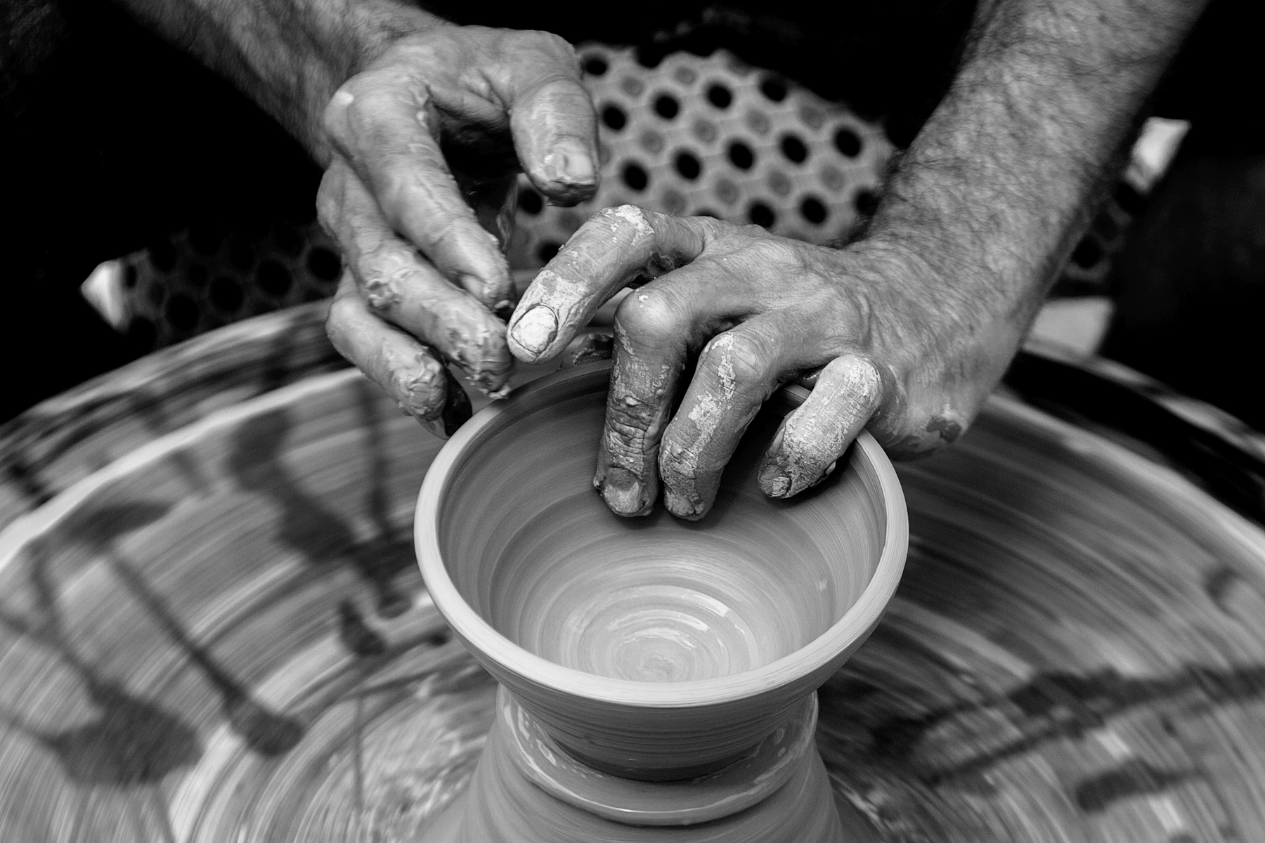 Clay-covered hands shape a small bowl on a spinning pottery wheel in a black-and-white photograph.