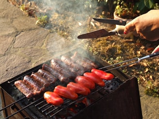 Grilled sausages and vibrant red bell peppers are sizzling on a barbecue under the sunlight. A hand is holding tongs, adjusting the items on the grill, with smoke rising gently.
