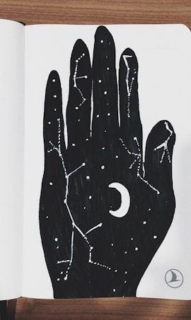 A stylized black hand drawing on white paper is filled with star constellations and a crescent moon, giving an impression of the night sky embedded within the hand. The art contrasts sharply against the wooden surface on which the notebook rests.