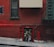 A red brick building features a narrow entrance labeled 'fire exit'. Two individuals are standing in the doorway, possibly engaging in a conversation or activity. A steep sidewalk slopes past the entrance, adding a sense of urban environment. A fire escape and barred window are visible on the building's facade.