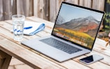 An outdoor shot of a laptop set up on a wooden table overlooking a serene mountain landscape.