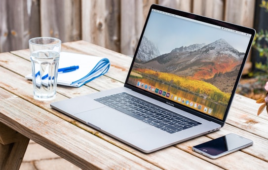 A silver laptop is open on a wooden table outdoors. The screen displays a mountain landscape wallpaper. Next to the laptop, there is a glass of water, a smartphone, and a notebook with a blue pen. The setting appears relaxed and casual, with natural lighting and a fence in the background.