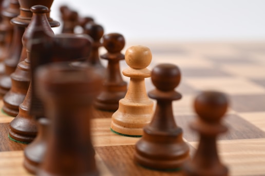 Close-up view of a chessboard featuring a wooden pawn in focus among other blurred chess pieces lined up diagonally. The board has a classic checkered pattern and the pieces have a glossy finish.