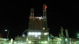 A large industrial facility illuminated at night, featuring a tall structure made of metal and glass with various lights and signals. The building appears to be part of a complex, with multiple pipes and machinery visible, some emitting steam or smoke.