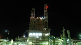 A large industrial facility illuminated at night, featuring a tall structure made of metal and glass with various lights and signals. The building appears to be part of a complex, with multiple pipes and machinery visible, some emitting steam or smoke.