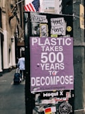 A street scene featuring a poster with purple background text that warns about the long decomposition time of plastic, stating 'Plastic takes 500 years to decompose'. The poster is affixed to a dark urban structure adorned with various stickers and graffiti. An American flag is visible in the background, and a person is seen walking away while pulling a suitcase.
