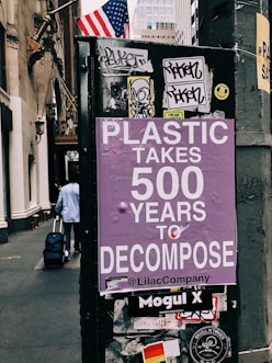 A street scene featuring a poster with purple background text that warns about the long decomposition time of plastic, stating 'Plastic takes 500 years to decompose'. The poster is affixed to a dark urban structure adorned with various stickers and graffiti. An American flag is visible in the background, and a person is seen walking away while pulling a suitcase.