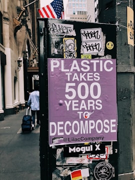 A street scene featuring a poster with purple background text that warns about the long decomposition time of plastic, stating 'Plastic takes 500 years to decompose'. The poster is affixed to a dark urban structure adorned with various stickers and graffiti. An American flag is visible in the background, and a person is seen walking away while pulling a suitcase.