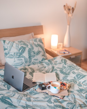 A cozy bedroom setting features a bed with green leafy-patterned bedding. On the bed, an open laptop and a book are accompanied by a tray holding a white mug and some snacks. A wooden bedside table holds a lamp, glasses, and a light decorative vase with dried plants. The lighting creates a warm and inviting atmosphere.