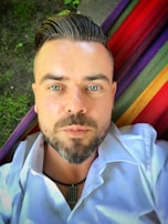 A man with a neatly groomed beard and styled hair is lying on a multicolored striped hammock. His eyes are open, and he is wearing a white shirt with a necklace visible. The background includes some patches of grass.