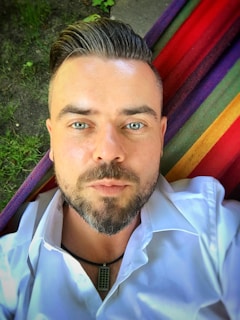 A man with a neatly groomed beard and styled hair is lying on a multicolored striped hammock. His eyes are open, and he is wearing a white shirt with a necklace visible. The background includes some patches of grass.