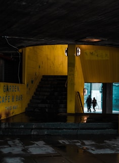 A dimly lit urban stairwell leads upwards, flanked by yellow walls with signage indicating directions to a garden, cafe, and bar. Two spherical lights illuminate the pathway. The ground appears wet, reflecting light. In the background, an opening leads to a brighter area where two silhouetted figures walk away.
