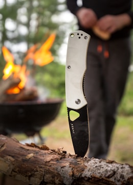 A knife with a white handle is embedded in a piece of wood, with a campfire burning in the background. A person is seen standing near the fire, slightly out of focus. The scene suggests an outdoor or camping setting with greenery visible.