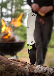 A knife with a white handle is embedded in a piece of wood, with a campfire burning in the background. A person is seen standing near the fire, slightly out of focus. The scene suggests an outdoor or camping setting with greenery visible.