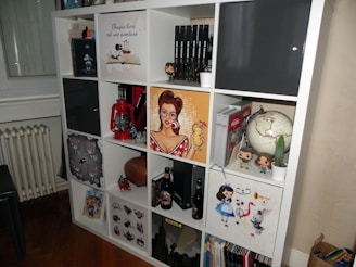 A multi-compartment shelf filled with various items including books, action figures, a globe, a red lantern, and decorative artworks. A pop art style poster of a woman holding a drink is prominently displayed, alongside Funko Pop figures and a small cactus in a white pot.