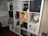 A multi-compartment shelf filled with various items including books, action figures, a globe, a red lantern, and decorative artworks. A pop art style poster of a woman holding a drink is prominently displayed, alongside Funko Pop figures and a small cactus in a white pot.