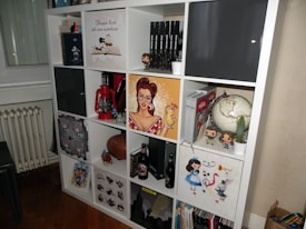A multi-compartment shelf filled with various items including books, action figures, a globe, a red lantern, and decorative artworks. A pop art style poster of a woman holding a drink is prominently displayed, alongside Funko Pop figures and a small cactus in a white pot.