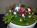 A decorative arrangement featuring small figurines resembling forest creatures, surrounded by colorful artificial plants and mushrooms on a grassy surface within a round container. The scene depicts a whimsical, miniature garden setting.