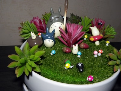 A decorative arrangement featuring small figurines resembling forest creatures, surrounded by colorful artificial plants and mushrooms on a grassy surface within a round container. The scene depicts a whimsical, miniature garden setting.