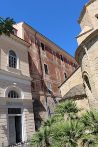 A historic architectural scene featuring a group of buildings with varied textures. The left building has light-colored stonework and an arched window above a doorway labeled 'Asilo Infantile'. To the right, an older, reddish-brown building with tall windows shows signs of aging. A round, stone structure with a textured roof is partially visible. Palm trees and a clear blue sky provide contrast and greenery.