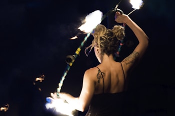 A woman with tattoos on her back is skillfully performing a fire dance. She is holding a flaming object, possibly a fire hoop or staff, in an artistic manner. Her hair is styled in buns, and she is wearing a black outfit, surrounded by darkness that highlights the flames and her figure.
