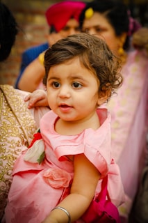 A young child wearing a pink dress with floral decorations is being held by an adult. The background includes several people, some wearing vibrant attire, suggesting a festive or celebratory event.