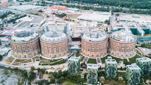 Four large, circular brick buildings with glass dome roofs are prominently displayed in an urban area. Construction cranes and modern buildings surround the historic structures, and greenery is visible in the background.