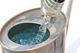 A close-up view of a metallic hose nozzle pouring clear water into a metal container. The water creates ripples and reflections within the container. The scene is brightly lit with minimal shadows.