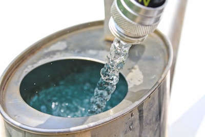 A close-up view of a metallic hose nozzle pouring clear water into a metal container. The water creates ripples and reflections within the container. The scene is brightly lit with minimal shadows.