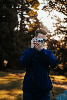 A person is holding a camera up to their face, capturing a photograph in an outdoor setting. The background features blurred trees and sunlight is filtering through, creating a serene atmosphere. The individual is wearing a dark jacket.