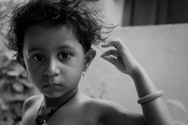 A young child with curly hair looks directly at the camera. The child is wearing a necklace and has an arm raised, touching their hair. The expression is serious and thoughtful, and the image is rendered in black and white, adding a timeless feel.