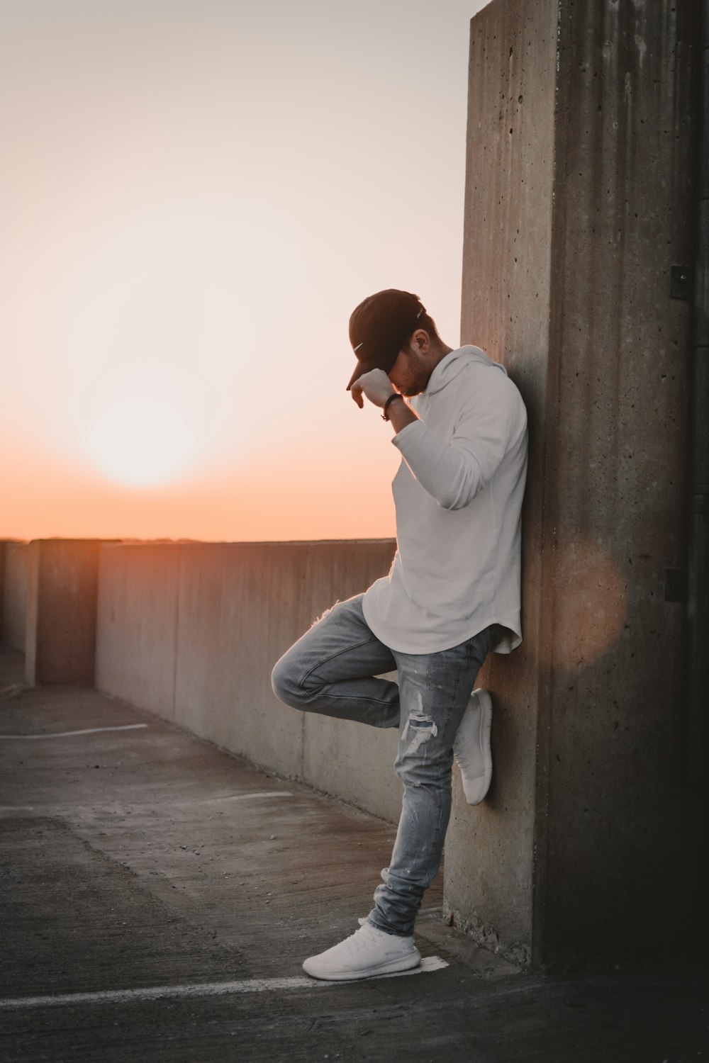 man in white jacket, black cap and blue distressed jeans leaning on gray concrete pillar column during orange sunset