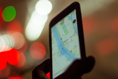 A smartphone being held up in one hand displays a map application showing a route in New York City. The screen is brightly illuminated against a blurred background filled with red, green, and white bokeh lights.