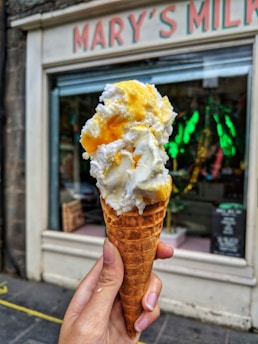 A hand is holding a waffle ice cream cone with a generous serving of white and yellow ice cream in front of a storefront. The storefront has 'MARY'S MILK' written at the top in red letters against a white background. The window displays various objects and green decorations.