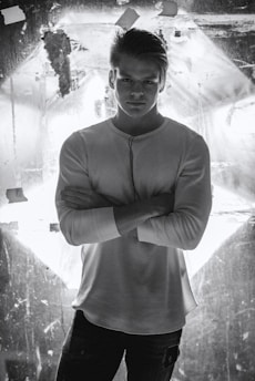 A man stands confidently with his arms crossed, wearing a white long-sleeve shirt. He is positioned against a backdrop that is illuminated, creating high contrast and dramatic lighting.