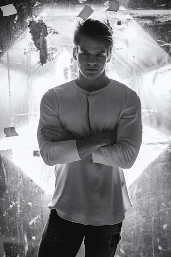 A man stands confidently with his arms crossed, wearing a white long-sleeve shirt. He is positioned against a backdrop that is illuminated, creating high contrast and dramatic lighting.