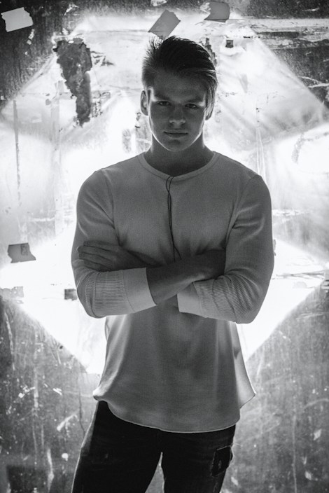A man stands confidently with his arms crossed, wearing a white long-sleeve shirt. He is positioned against a backdrop that is illuminated, creating high contrast and dramatic lighting.
