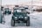 A rugged, dark green off-road vehicle with snow-covered tires is parked at a snowy gas station. Several people, wearing winter clothing, are gathered near other large vehicles in the background. The landscape features snow-covered hills under a clear sky.
