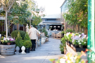 A quaint outdoor cafe setting surrounded by lush greenery and potted plants. There are various types of flowers on display, along with outdoor seating arrangements. A person wearing a beige coat and black boots is seen walking towards the seating area. The atmosphere is calm and inviting, with a rustic and natural ambiance.