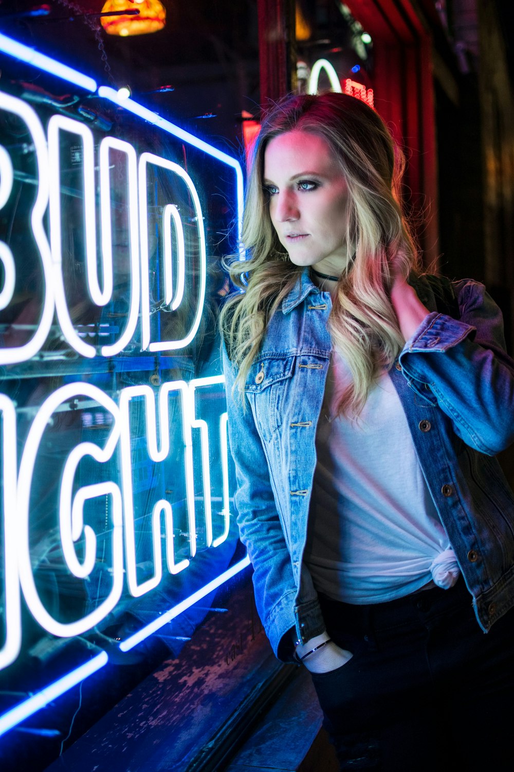 woman wearing white shirt and blue denim button-up jacket near Bud Light neon signage