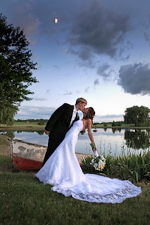 A newlywed couple shares a romantic kiss near a tranquil lake, surrounded by lush greenery. The bride, in a white gown, holds a bouquet of flowers, while the groom wears a black tuxedo. A small, rustic boat sits nearby, and the sky above features scattered clouds with a visible moon.