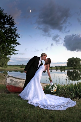 A newlywed couple shares a romantic kiss near a tranquil lake, surrounded by lush greenery. The bride, in a white gown, holds a bouquet of flowers, while the groom wears a black tuxedo. A small, rustic boat sits nearby, and the sky above features scattered clouds with a visible moon.
