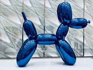 A large, shiny blue sculpture resembling a balloon animal in the shape of a dog stands prominently in a modern gallery space. The background consists of geometric patterns and reflected light, creating a sense of depth.